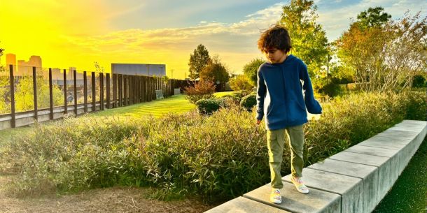 Child walking a low wall in a neighborhood park at sunset, city skyline glowing in the background, capturing the everyday rhythm you get from a short‑term rental stay.